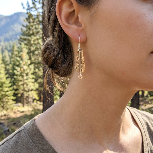 Model outdoors wearing Mokume-Gane sway earrings with Oregon Sunstone, complemented by a sunstone ring, with detailed jewelry visible against scenic background.