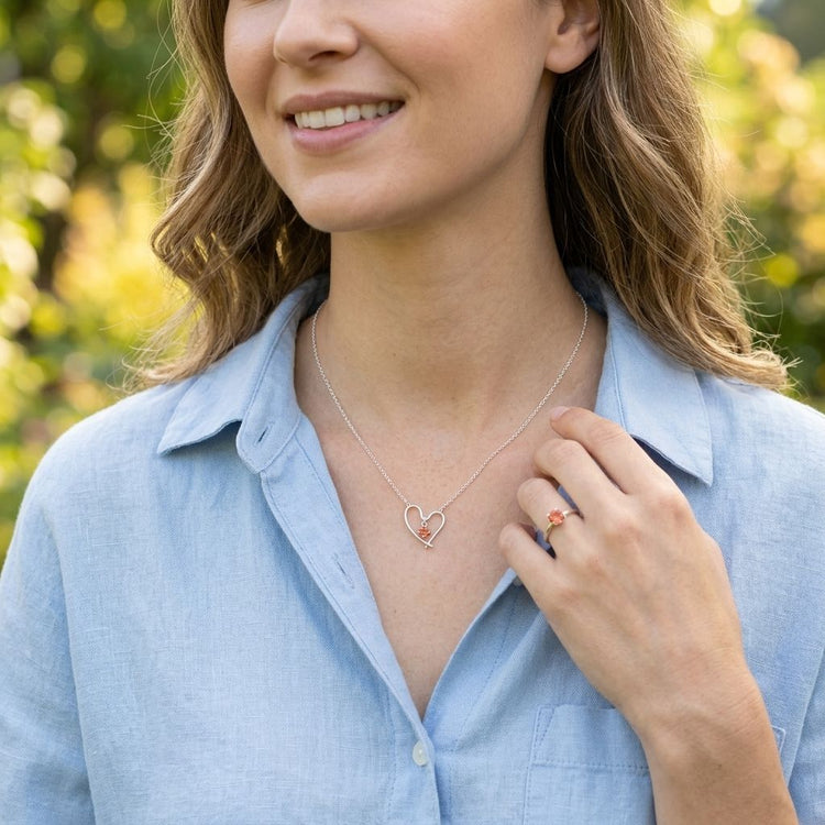 Model in casual outdoor setting wearing Valentine Heart Necklace with Oregon Sunstone, smiling and showing her hand.