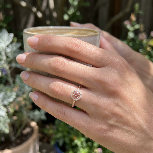 High-resolution close-up of small white gold sunstone engagement ring with diamond halo, set against a natural outdoor backdrop with soft natural lighting.