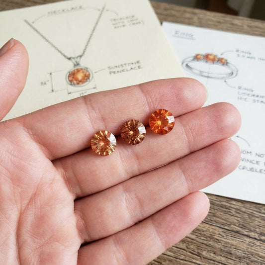 Highly detailed close-up of three fiery orange Oregon Sunstones, 9mm, held in a model's hand with natural background, showcasing brilliance and clarity.