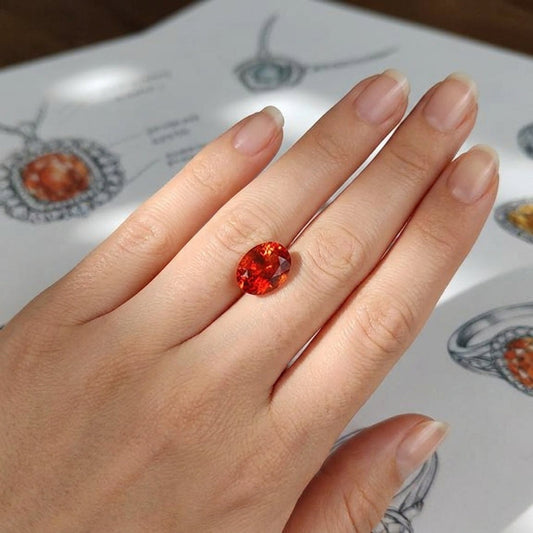 High-resolution close-up of a small oval Oregon Sunstone gemstone with fiery copper inclusions, resting on a flawless hand with jewelry sketches in background.