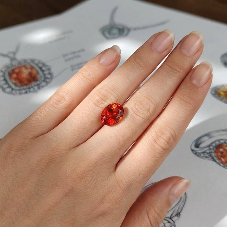 High-resolution close-up of a small oval Oregon Sunstone gemstone with fiery copper inclusions, resting on a flawless hand with jewelry sketches in background.