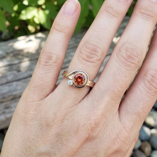 Close-up of an elegant hand wearing a delicate Oregon Sunstone ring with diamond accent, set outdoors with soft natural lighting.