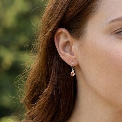 Female model wearing sterling silver hoop earrings with peach-hued Oregon Sunstone dangles, outdoors with natural scenery and relaxed pose.