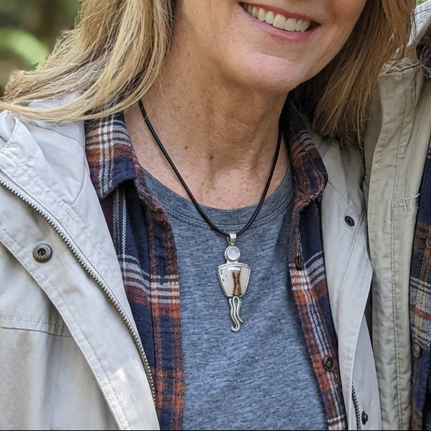 Close-up of a hand holding a polished gem ring with vibrant color, subtle necklace peeking from collarbone on a blurred outdoor background.