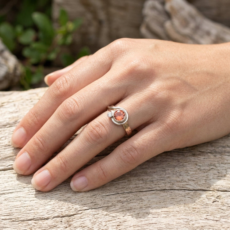 Close-up of a hand gently holding or resting on a textured wood surface, showcasing a vibrant orange Sunstone ring with warm lighting.