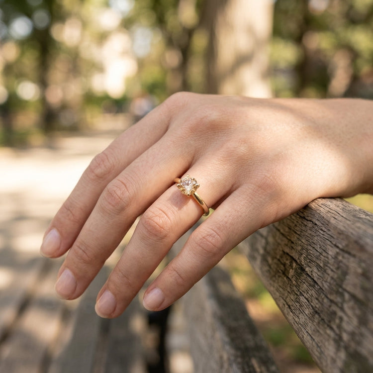 Casual outdoor scene of a model on a park bench showcasing a tiny rose gold Oregon Sunstone engagement ring in dappled sunlight.