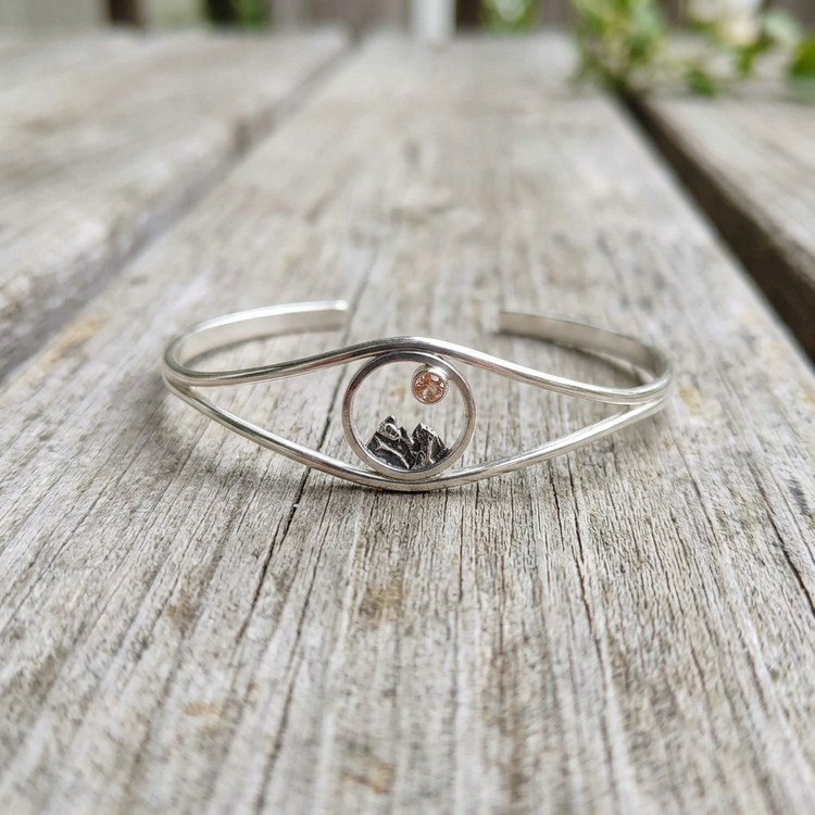 Artistic shot of Mini Mountain Cuff Bracelet placed on rustic wooden outdoor table, highlighting oxidized details, polished silver, and warm Sunstone glow.