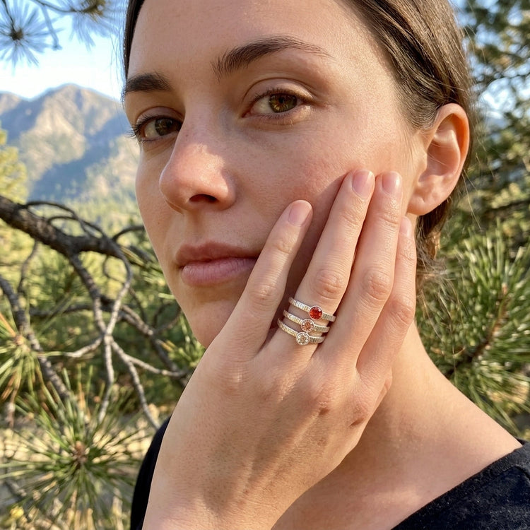 Portrait of a model with hands near her face, showcasing small sunstone stacking rings against a scenic pine and mountain backdrop.