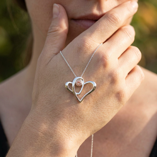Artistic shot of sterling silver 'Two Hearts as One' necklace on a model's hand, showing micro-details of the Sunstone in a relaxed outdoor setting with natural lighting.