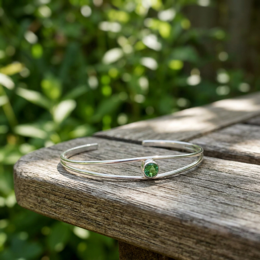 Elegant silver split cuff bracelet featuring a vibrant green sunstone, showcased on rustic wood amid a lush green garden with soft natural light.