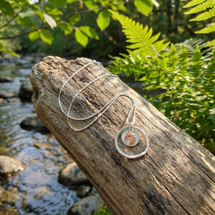 Sunstar necklace draped over driftwood with flowing water and greenery in the background, highlighting natural colors and peaceful outdoor setting.
