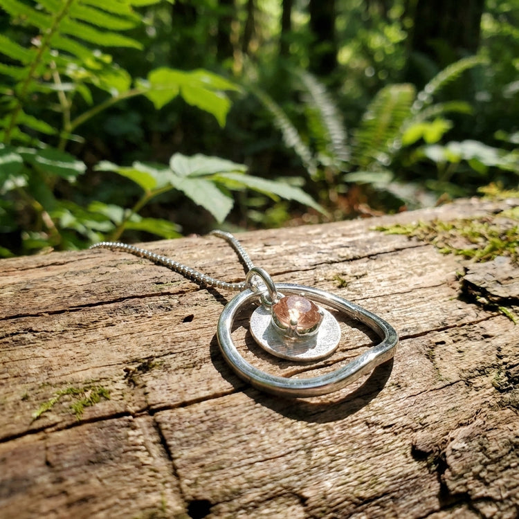 Delicate Sunstar necklace with 5mm Peach Oregon Sunstone pendant on wooden surface with green foliage and sunlight filtering through leaves.