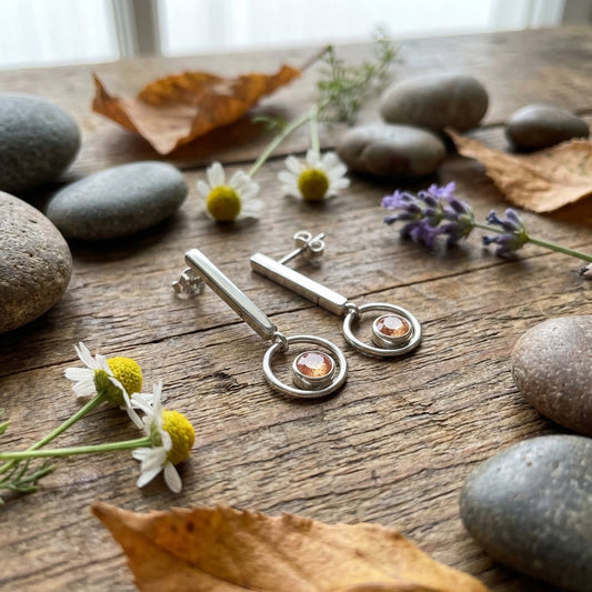 Artistic flat-lay of Sunstone Modern Dangle Earring on wooden surface with pebbles, leaves, and wildflowers, emphasizing its sparkle and design.
