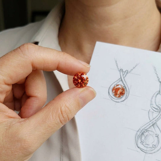 Close-up of a 10mm vivid orange Oregon Sunstone held between fingers, highlighting its fiery sparkle and clarity with a blurred jewelry sketch background.
