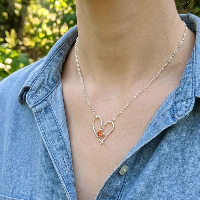 Female model wearing Valentine Heart Necklace with Oregon Sunstone, outdoors with greenery, showcasing delicate jewelry on her neck.