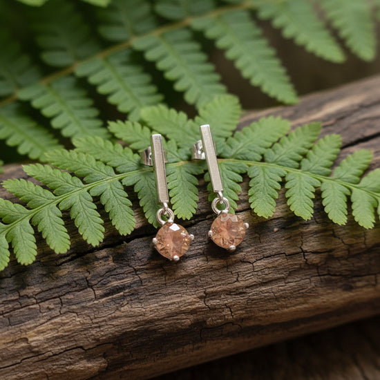 A pair of silver-toned dangle earrings with peach-colored faceted gemstones, displayed on a textured gray stone stand against a plain white background.