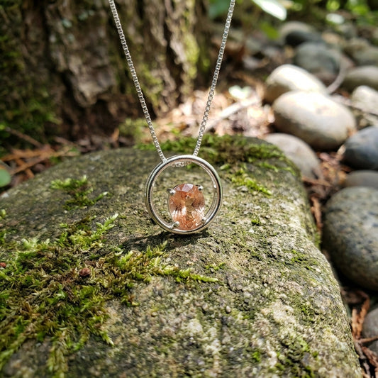 Artistic shot of Silver Oregon Sunstone Eclipse Necklace hanging from a rustic branch, emphasizing iridescent copper schiller and intricate facets against natural textured background.