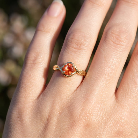 Delicate hand showcasing a fine gold Sunstone engagement ring with sunset hues, against a softly blurred natural background emphasizing craftsmanship.