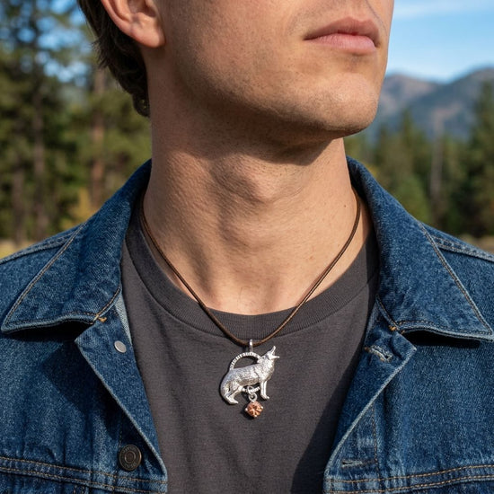 Close-up of the Howling Wolf Pendant hanging from a rugged leather cord on a male model's neck, set against a natural landscape background with sunlight highlighting silver details.