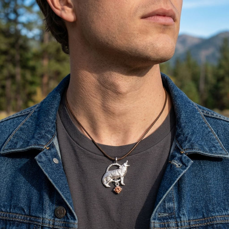 Close-up of the Howling Wolf Pendant hanging from a rugged leather cord on a male model's neck, set against a natural landscape background with sunlight highlighting silver details.