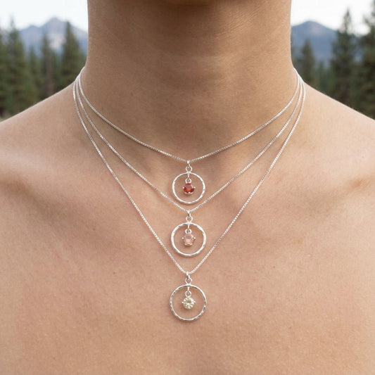 Detailed view of tiny Oregon Sunstone Saturn Pendant lying on rustic wood with blurred mountain and pine background, showcasing micro-texture and fiery gem sparkle.