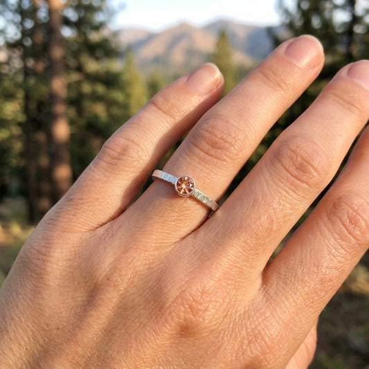 Close-up of a sterling silver sunstone ring with textured bezel, set on a natural hand against a scenic mountain landscape.