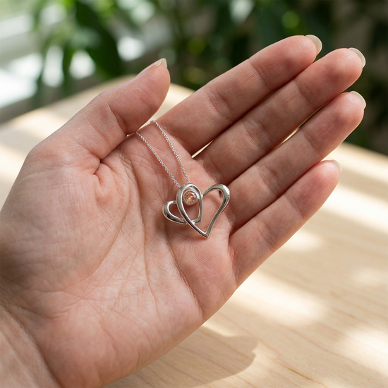 Close-up of sterling silver 'Two Hearts as One' necklace with Oregon Sunstone showing shimmer and micro-texture on a manicured hand in outdoor natural setting.