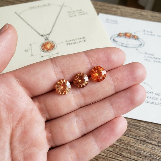 Highly detailed close-up of three fiery orange Oregon Sunstones, 9mm, held in a model's hand with natural background, showcasing brilliance and clarity.