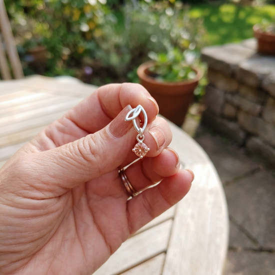 Relaxed hand holding sterling silver Twinleaf earring with Oregon Sunstone in a cozy natural outdoor scene, with soft shadows and blurred background.