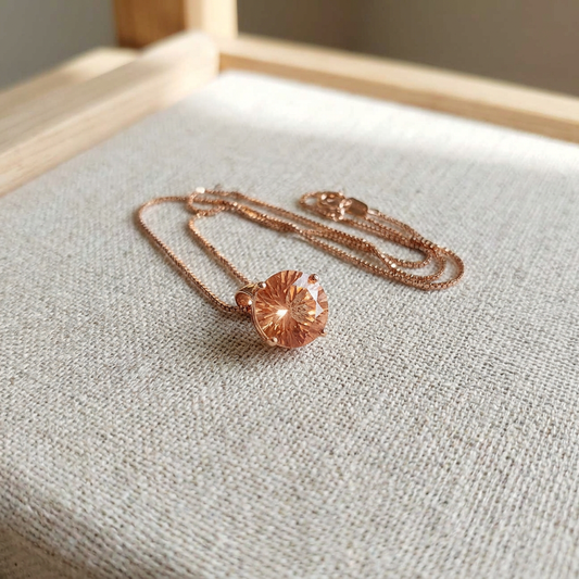 Close-up of a 9.5mm Oregon Sunstone pendant set in 14K rose gold, showcasing fiery brilliance and intricate facets on a soft wooden surface.