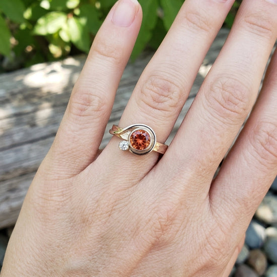 Close-up of an elegant hand wearing a delicate Oregon Sunstone ring with diamond accent, set outdoors with soft natural lighting.