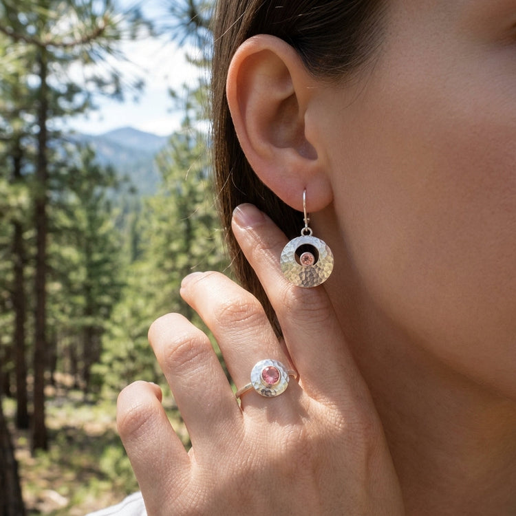 Close-up of textured silver Galilean Moon earrings with peach Oregon Sunstone on a hand against a pine forest background.