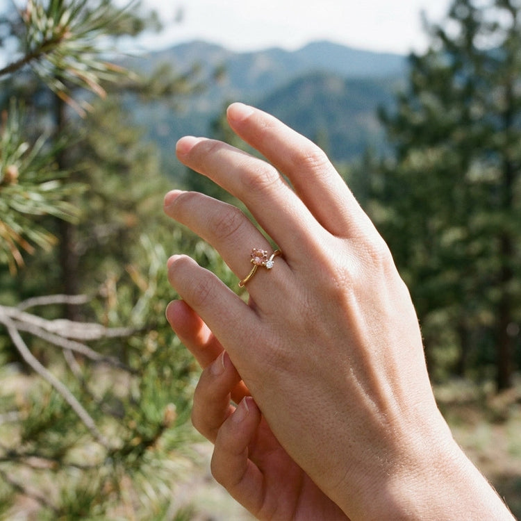 Full-body shot of a woman outdoors, showing the 10K gold Sunstone ring on her finger, with a backdrop of pine trees and mountains, holding wildflowers.