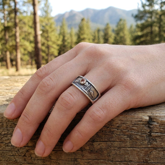 Full-body shot of a model outdoors wearing the sterling silver Three Peaks and Ponderosa Mountain ring on a relaxed hand, with mountains and pine trees.