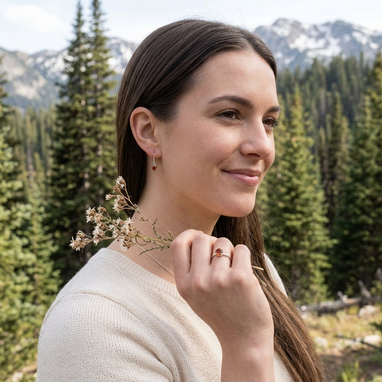 Full-body outdoor shot of a model wearing tiny silver sunstone earrings with a matching ring, in a natural setting with pine trees and mountains, emphasizing jewelry scale and style.