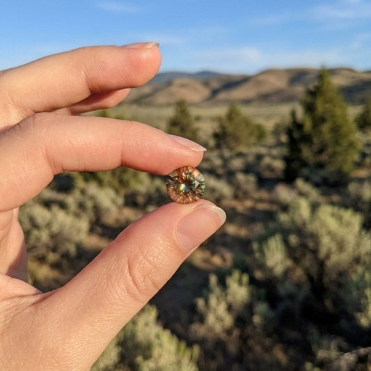 Detailed view of a small bi-color Oregon Sunstone held by a flawless hand, set against a soft-blurred desert environment, showcasing internal sparkles and color shifts.