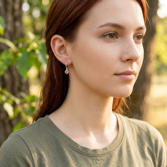 Close-up of hand holding ring with bright gemstone near face, ear visible with tiny silver hoop earring with peach sunstone, outdoors.