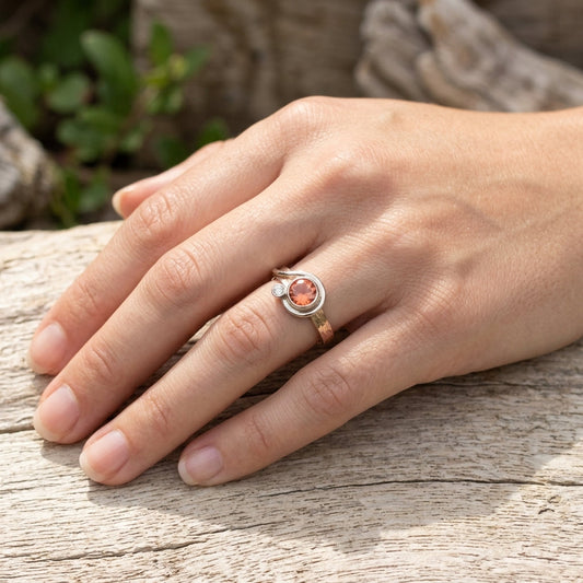 Close-up of a hand gently holding or resting on a textured wood surface, showcasing a vibrant orange Sunstone ring with warm lighting.