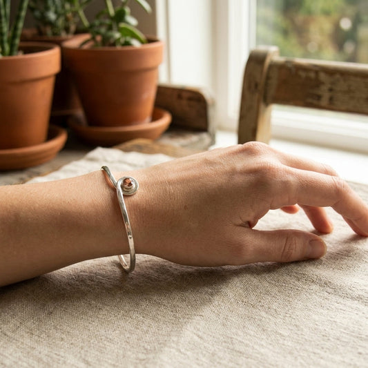 Side view of a hand with a silver Oregon Sunstone bracelet, resting on linen fabric in a bright indoor setting, showcasing bracelet texture and gemstone hues.