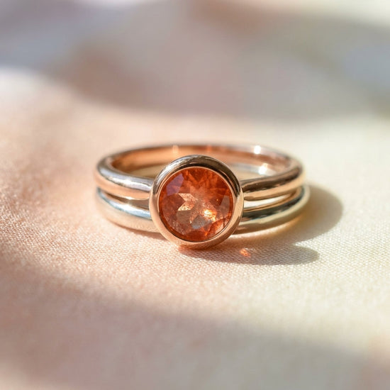 Close-up macro of a sunstone bezel ring against pastel fabric, highlighting the craftsmanship, fiery facets, and smooth finish in natural light.