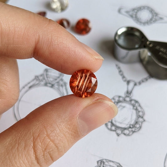 Macro shot of a small Oregon Sunstone gem with red-orange hue and shimmer, held in a hand with blurred jewelry sketches in the background.
