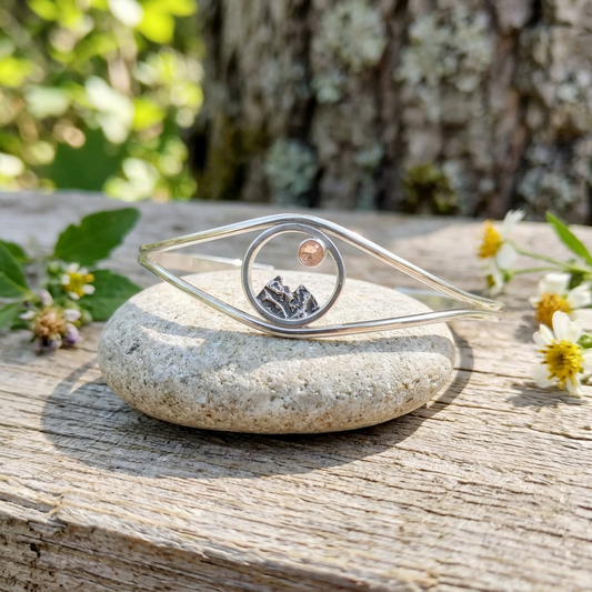 Close-up of sterling silver Mini Mountain Cuff Bracelet with Oregon Sunstone, detailed mountain engraving, natural outdoor background with greenery and wood textures.