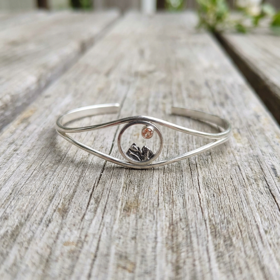 Artistic shot of Mini Mountain Cuff Bracelet placed on rustic wooden outdoor table, highlighting oxidized details, polished silver, and warm Sunstone glow.