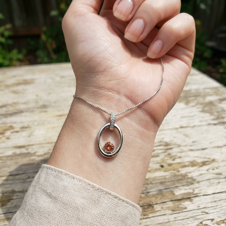 Close-up of a hand with a tiny silver Oregon Sunstone pendant on the wrist, illuminated by natural light, in a casual setting.