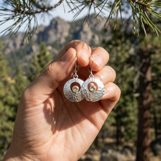Hand holding textured silver Galilean Moon earrings with champagne Oregon Sunstone in an outdoor pine and mountain setting.