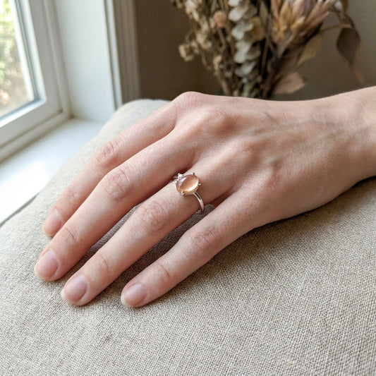 Delicate hand wearing Odyssey Sunstone Cabochon Ring with tiny diamond, against a neutral fabric background, highlighting the warm tones and craftsmanship.