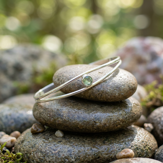 Silver split cuff bracelet with green sunstone draped over smooth river stones, captured outdoors with natural daylight and greenery.