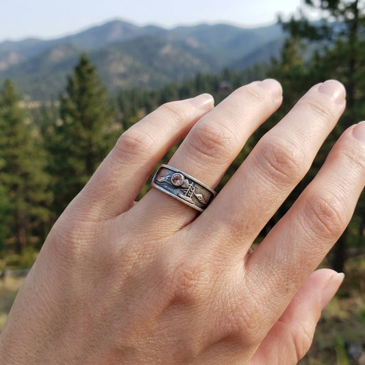 Model’s hand with ring finger wearing sterling silver Mountain Band, in a soft outdoor landscape with pine trees and mountains in background.