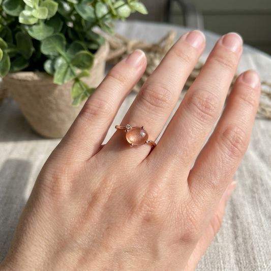 Close-up of a delicate rose gold ring with peach Sunstone cabochon and diamond accent on a model's hand, natural outdoor light.